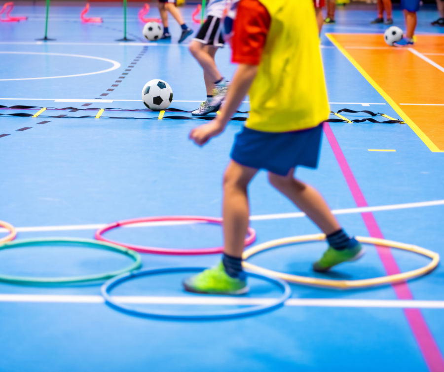 Kind in sportkleding loopt door twee gekleurde ringen die op een blauwe gymzaal liggen. Op de achtergrond ligt een witte voetbal met zwarte blokken.