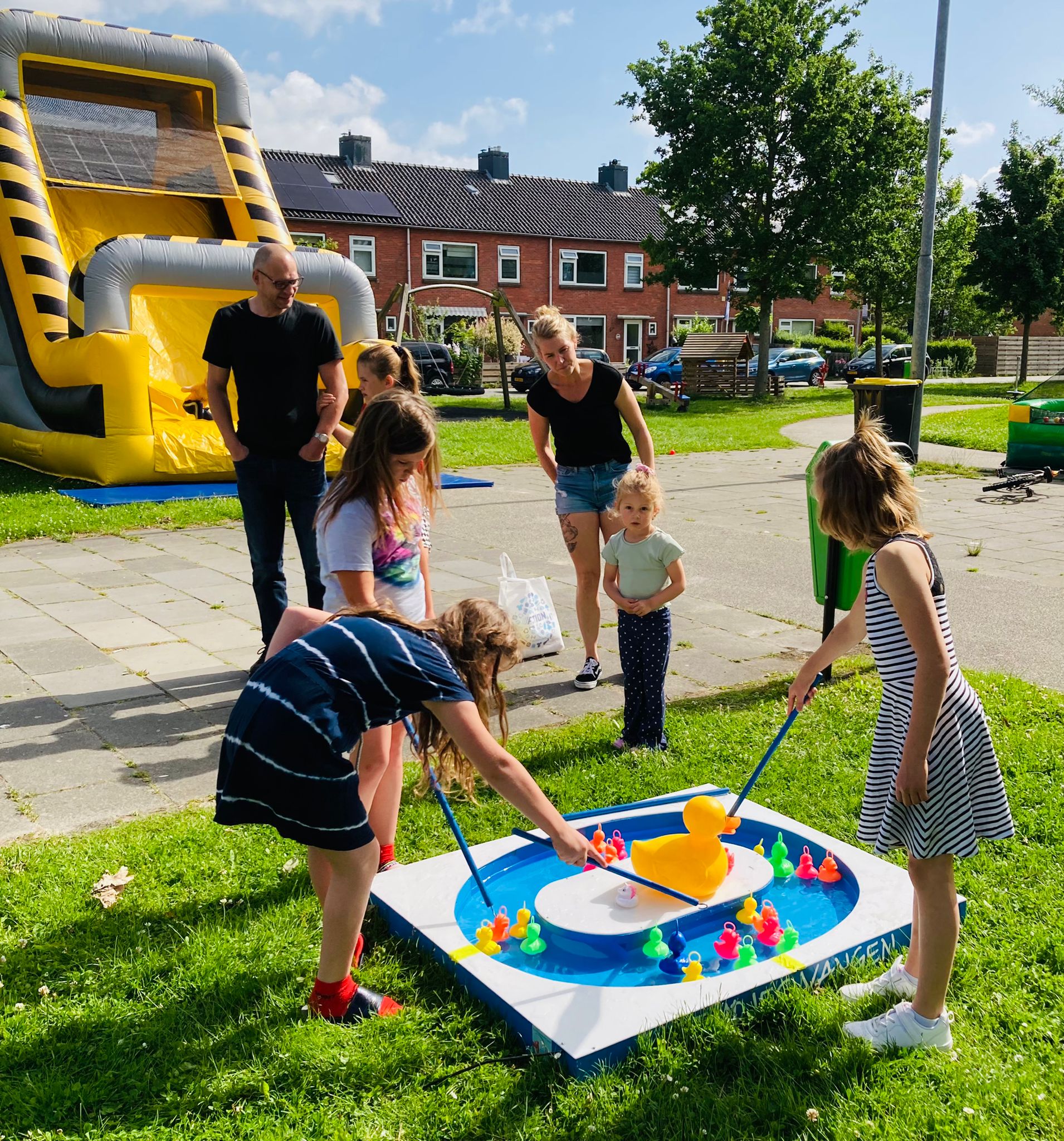 Op de achtergrond staat een geel springkussen. Op de voorgrond spelen 2 kinderen. Ze proberen met een stok gekleurde badeenden uit water te vissen. Daaromheen kijken ouders en kinderen.