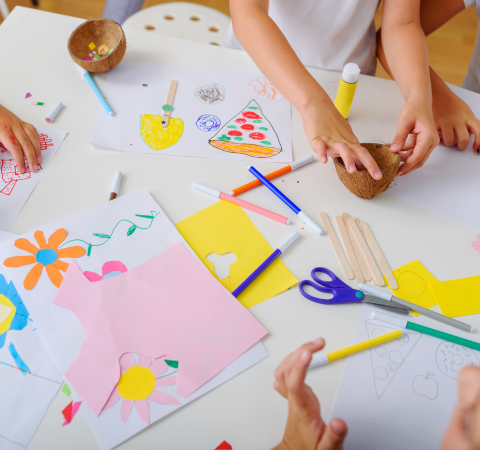 Witte tafel gevuld met verschillende gekleurde papieren. Er ligt een schaar en stiften. Handen van kinderen zijn aan het knutselen