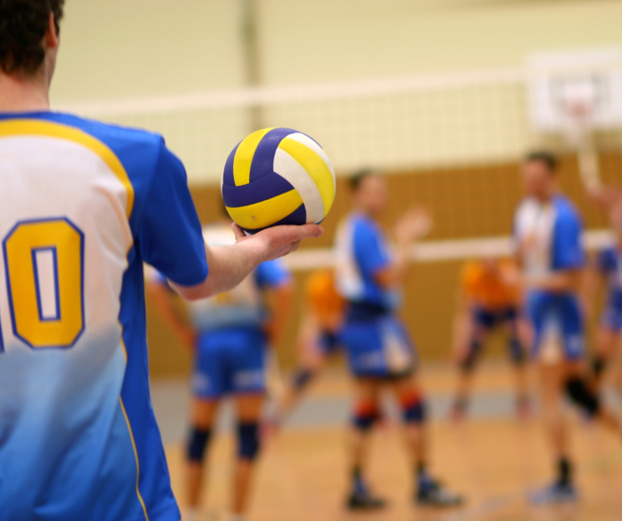 Man met een blauw sportshirt heeft een volleybal in zijn rechterhand. Hij staat klaar om op te slaan. Voor hem staat zijn team voor een volleybal net. 