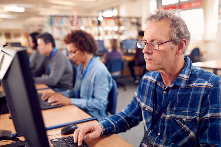 Een man met grijs haar, een bril en een blauwe blouse zit achter een computer. Naast hem zit een dame ook achter een computer. Beide ogen een leeftijd te hebben van 60 plus.