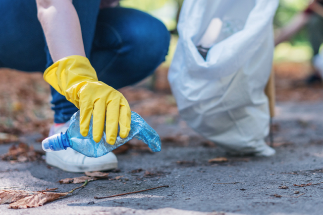 Hand met een gele handschoen pakt een plastic water flesje op. Persoon zit gehurkt met een witte vuilniszak.