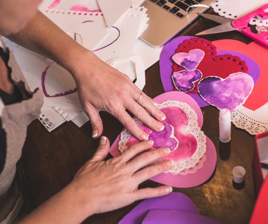 Twee handen plakken een paars hartje op een groter roze hartje van papier. eromheen liggen allerlei paarse, rode en roze gekleurde hartjes van papier.